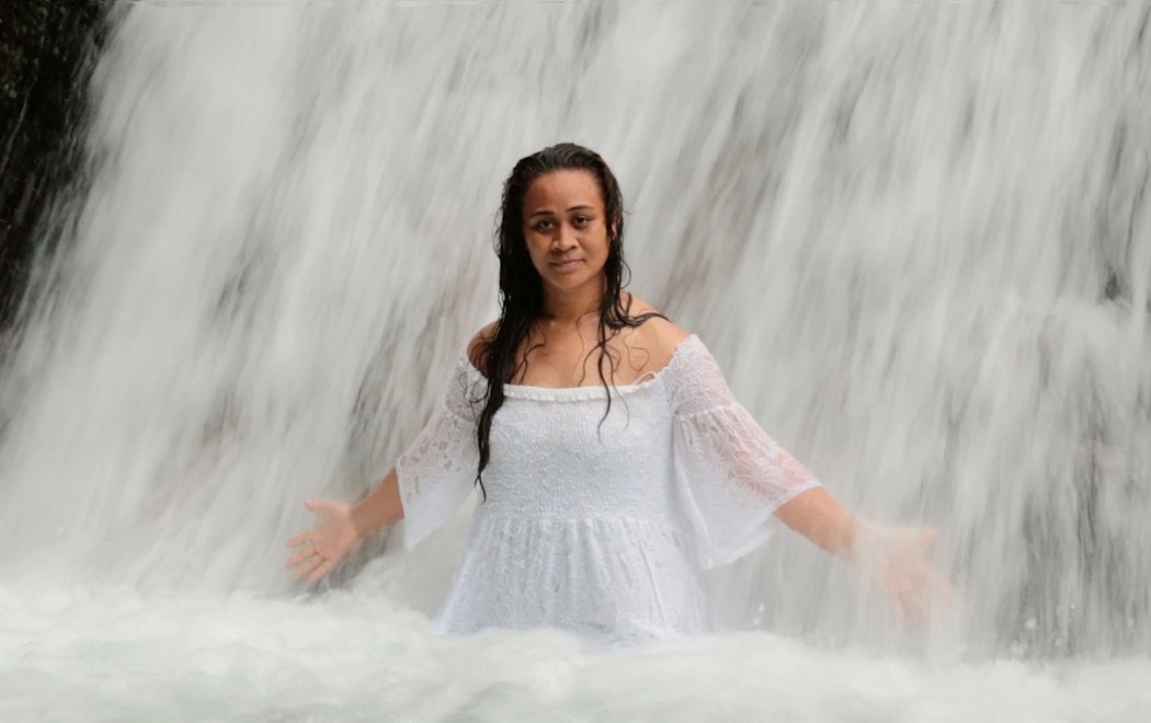 A woman in a white dress stands with her arms open under a waterfall. The white water is cascading over her, she looks serious but calm